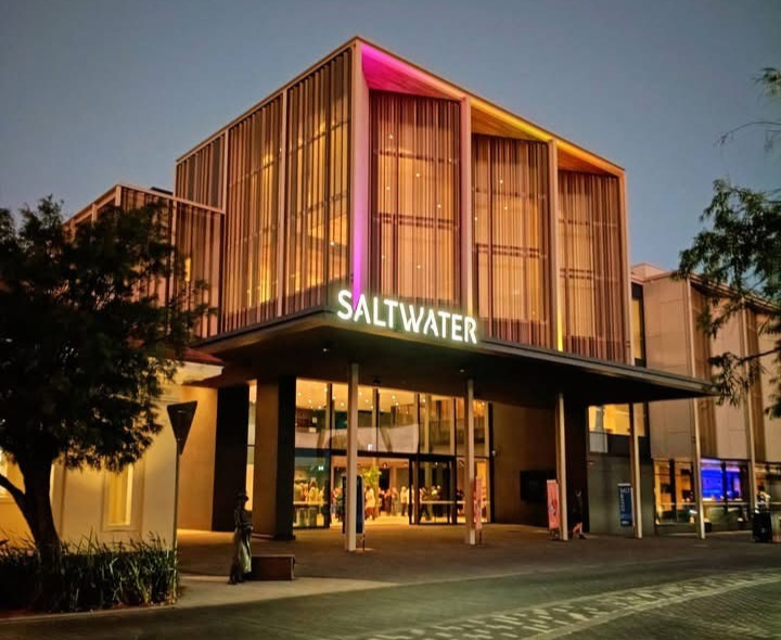 Photo of Saltwater Busselton building, taken from across street. It is nighttime and the building lights are on, casting a pink and yellow hue over the front of the building.
