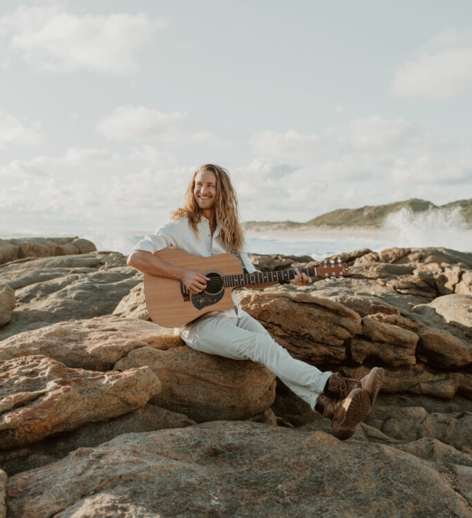 Man playing guitar, sitting on rocks by the ocean.